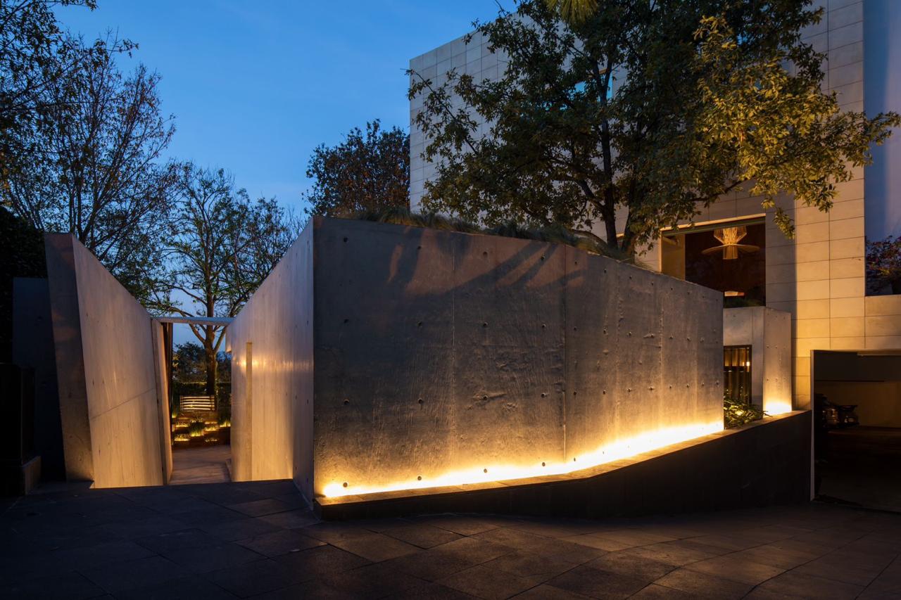 Esta imagen muestra un Pabellón MF, una ampliación independiente a una vivienda unifamiliar existente. El pabellón está iluminado por la noche, lo que crea un ambiente cálido y acogedor. La fachada del pabellón está hecha de concreto aparente, lo que le da un aspecto moderno y minimalista.