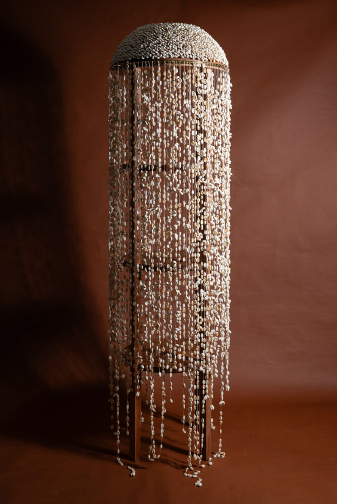 The ‘Orí’ shelf is a tubular shelf made from pink Jequitibá, brass details and more than seven thousand conch shells that adorn the head. Being a hollowed-out shelf, its closure is light and fluid, created by the volume of the strings of cowries, which both hide what's inside the Orí and allow what's inside to breathe.