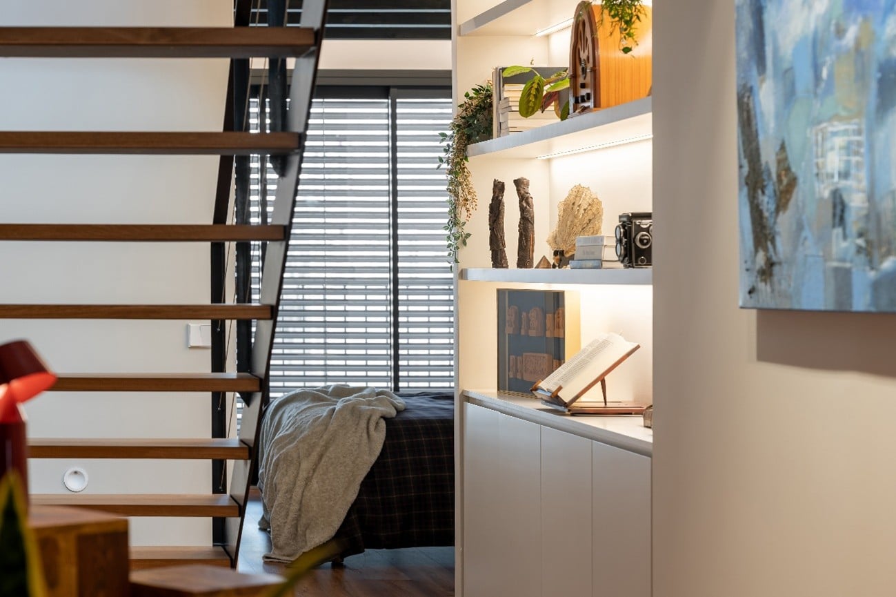 escalera de dúplex con librería retroiluminada blanca y al fondo pie de cama