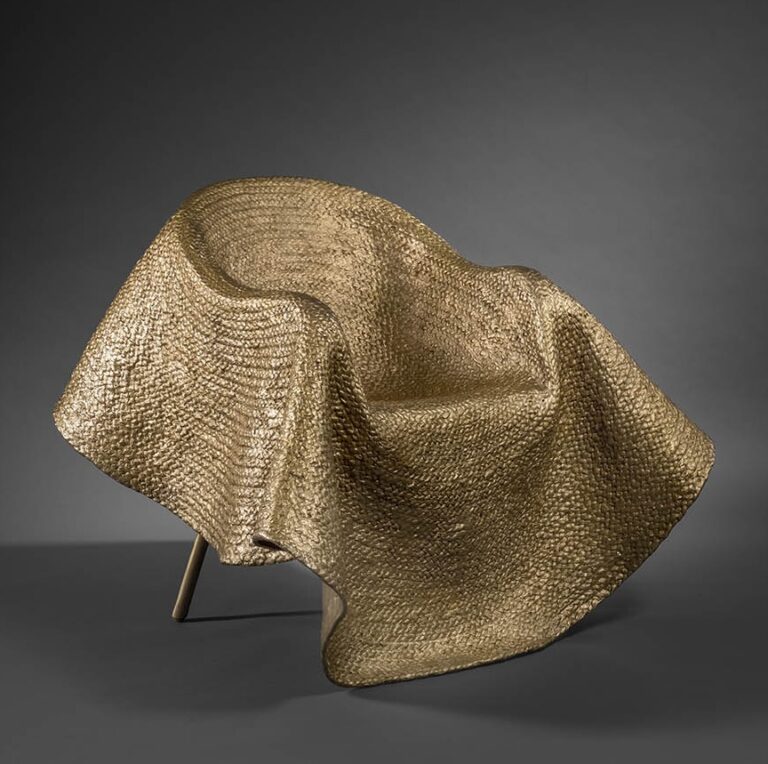De oro parece. Este sillón escultural forma parte de la serie que el diseñador de moda italiano, Stefano Pilati, ha realizado para la lujosa compañía francesa Pinto. No es de extrañar que quien fue miembro de la dirección creativa de Yves Saint Laurent, Prada o Cerruti ahora nos sorprenda con su implicación en el mundo del mueble. Pilati desarrolla este trabajo a partir de una alfombra de bronce con apariencia de fibras naturales, construida mediante la técnica de fundición a la cera perdida y cosida sobre un sillón de mimbre. La inspiración y su envolvente de gran tamaño vienen del drapeado y otras siluetas que se le ocurrieron cuando cubrió algunos enseres viejos con cojines, paja y yute en su casa de Sicilia. “Aunque no es lo mismo diseñar una prenda que diseñar mobiliario”, nos dice, “hay afinidades con el aspecto industrial del diseño de moda, pues también en él se hace necesario recordar la física, las proporciones y las tensiones compartidas entre las partes”. De ahí que su obra se erija como una escultura muy couture en desafío con la gravedad.